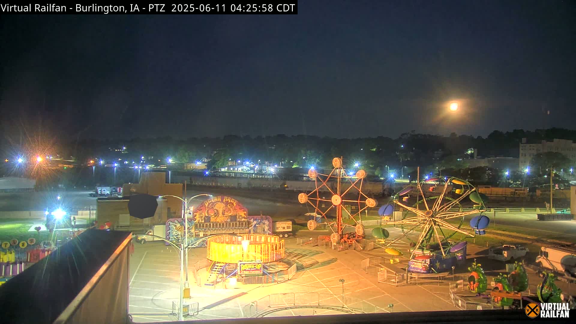 A nighttime view of a carnival midway with several illuminated rides under a bright moon.