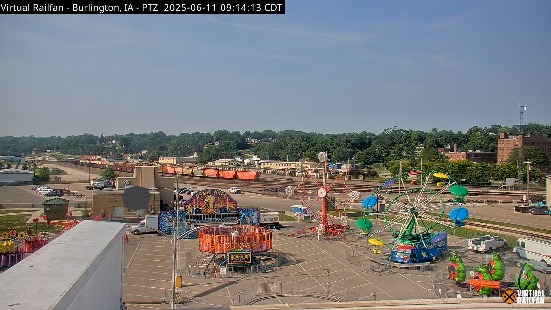 Under a clear sky, a carnival with several rides sits in a parking lot next to a train yard and a line of boxcars.
