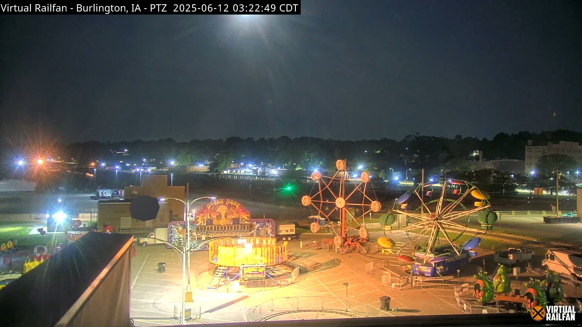 A nighttime view of a carnival midway with several illuminated rides and games, under a mostly clear sky.