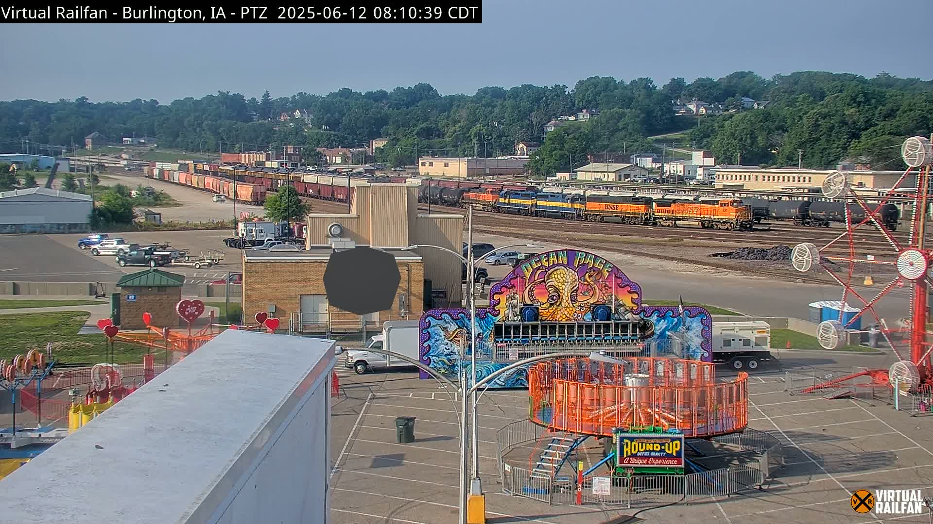 A long freight train sits in a rail yard next to a carnival midway under a clear, sunny sky.