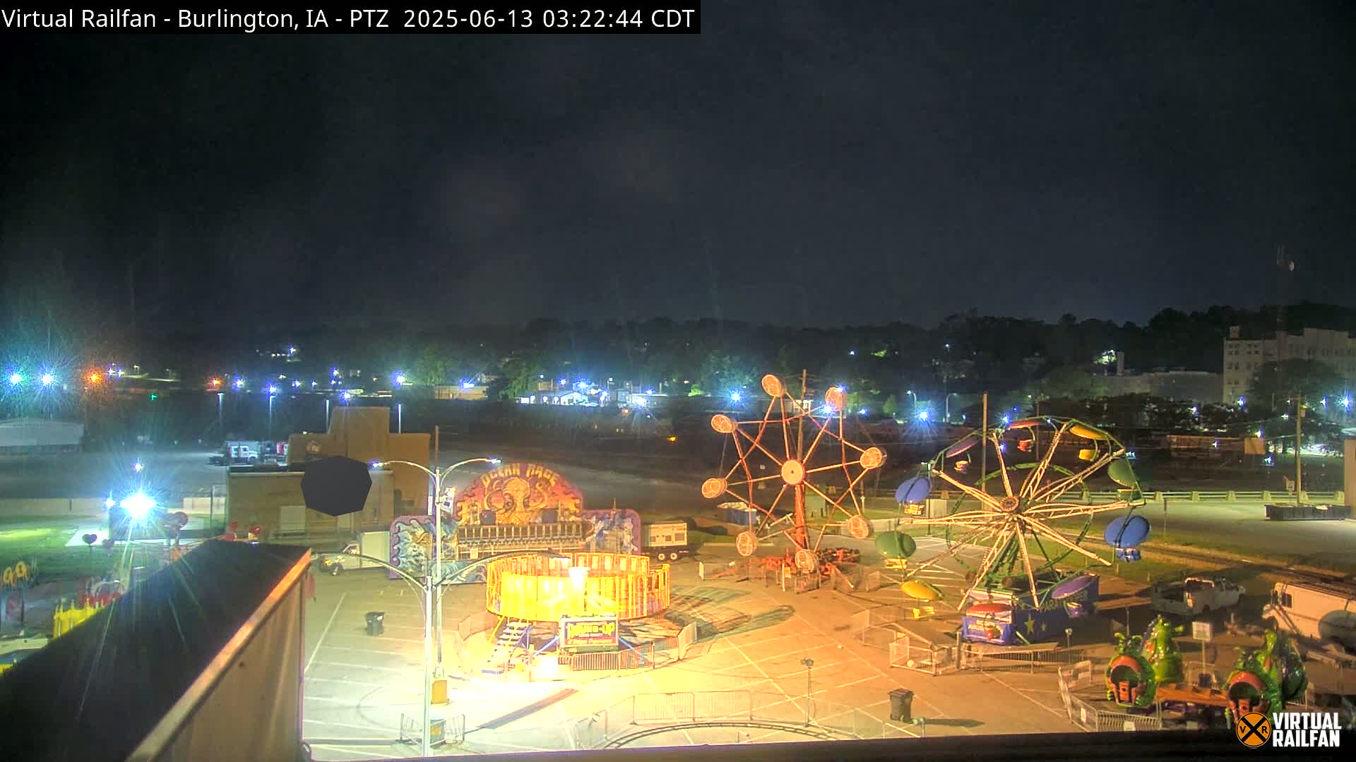 At night, a carnival midway with several illuminated rides is visible, situated in a parking lot next to a town under a clear, dark sky.
