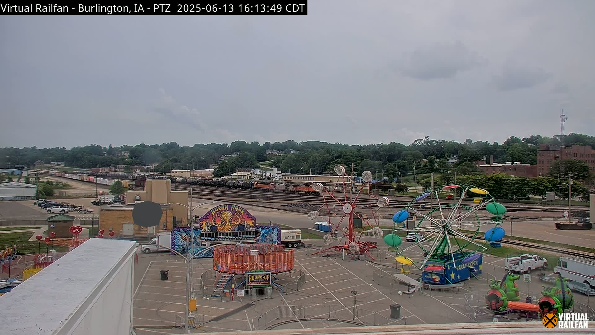 A carnival with several rides sits in a parking lot next to a railroad yard under an overcast sky.