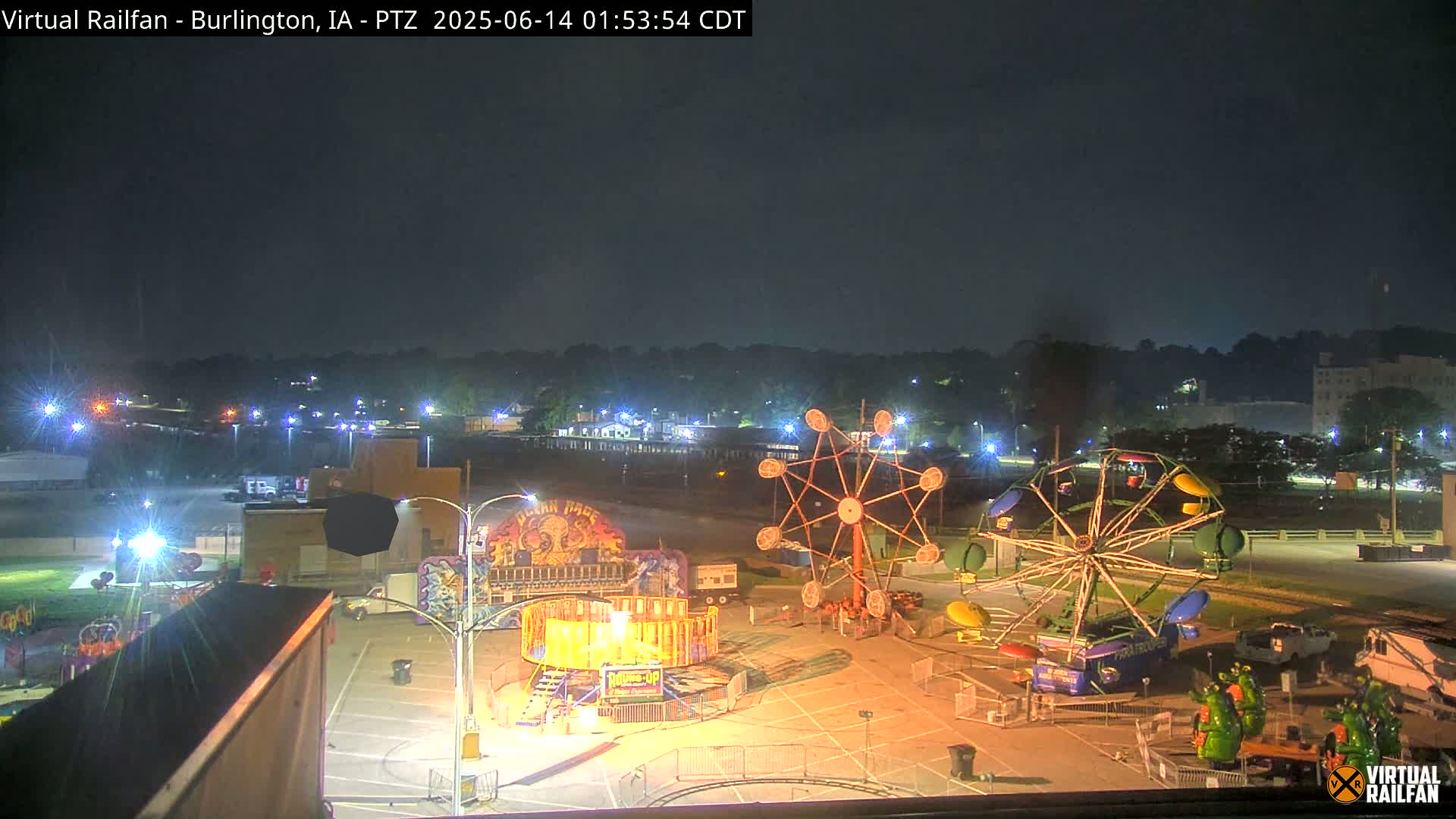 A carnival with several rides sits in a parking lot next to a railroad yard under an overcast sky.