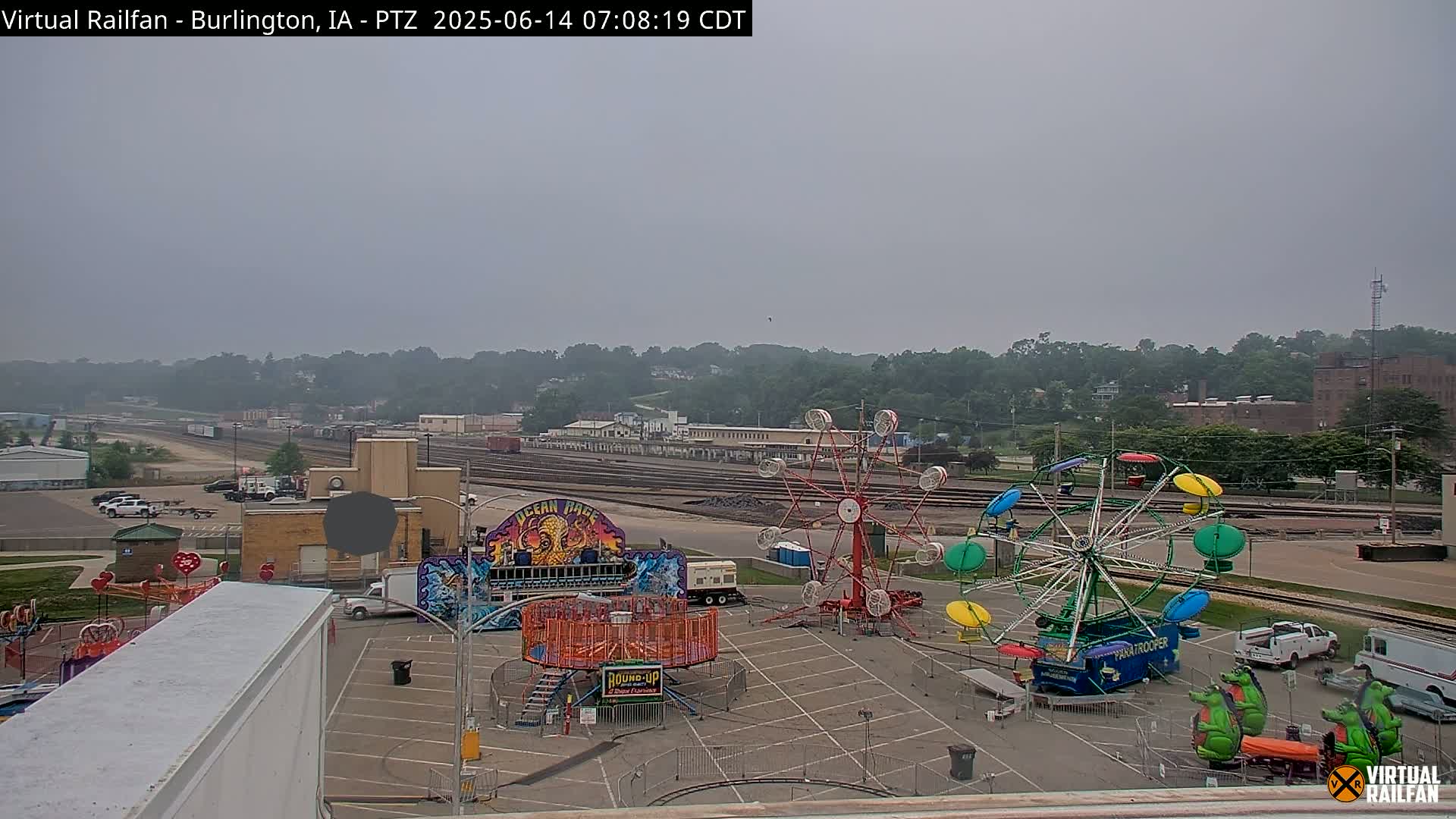 A fairground with several carnival rides sits in a parking lot next to railroad tracks, all under a hazy, overcast sky.