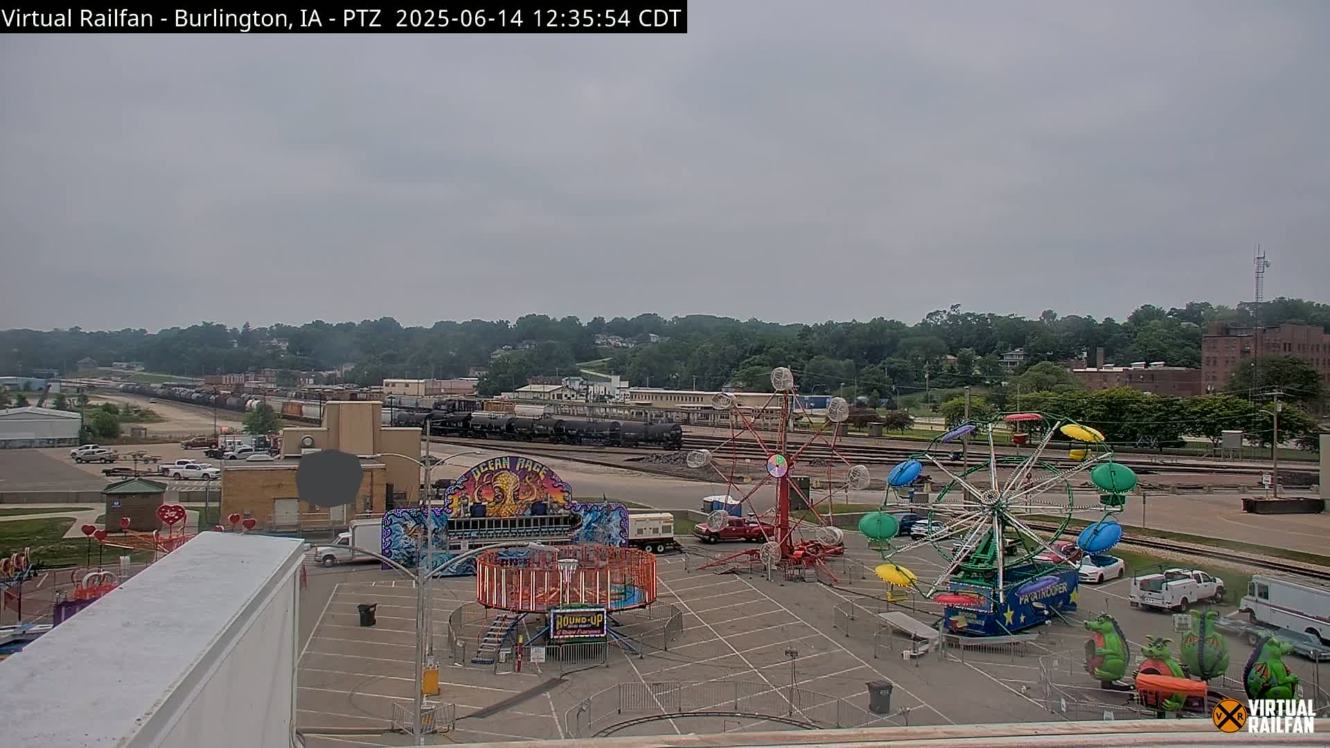 A carnival midway with several rides is situated in a parking lot next to railroad tracks and a line of freight cars under an overcast sky.