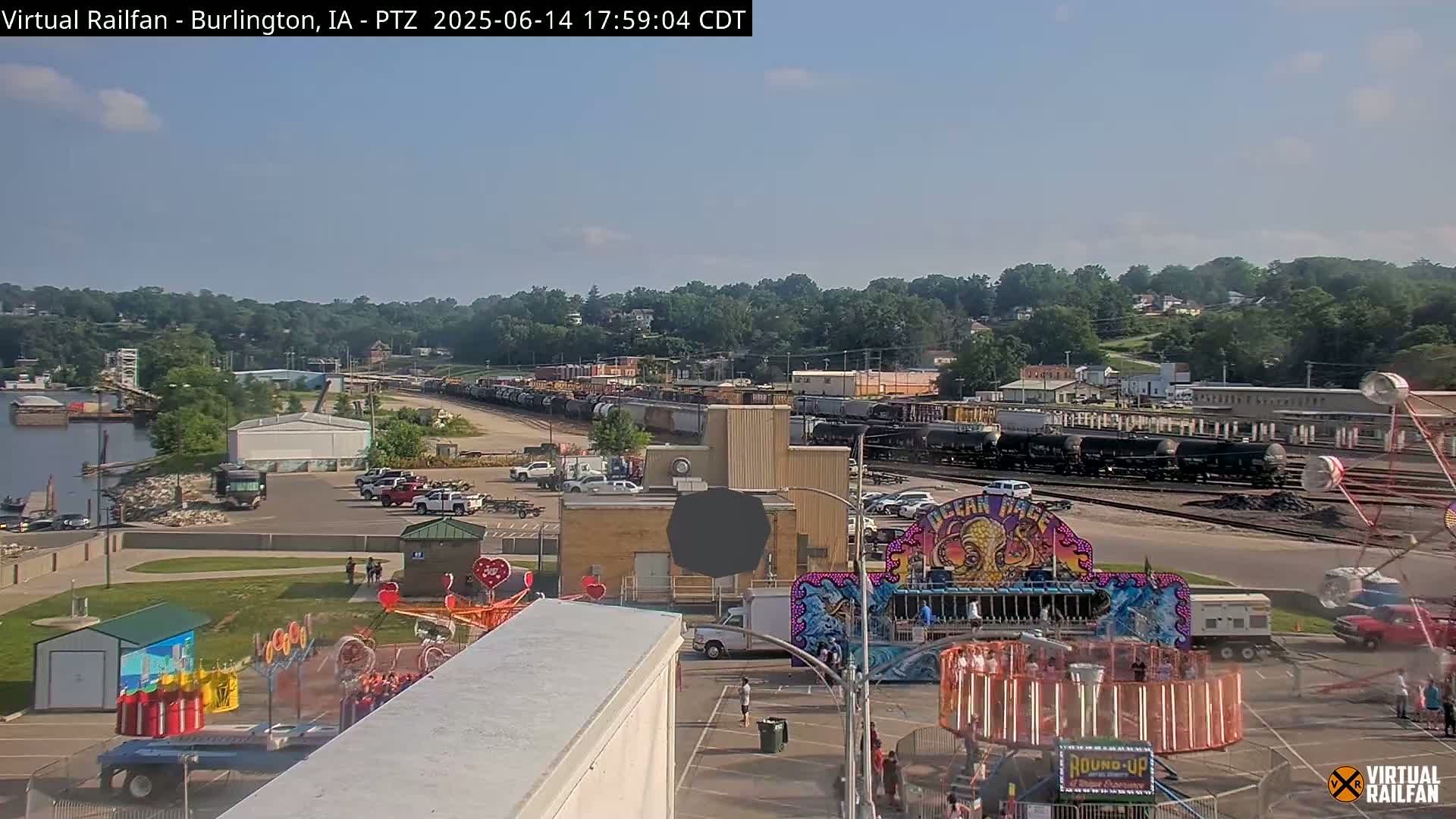 A carnival midway with several rides is situated in a parking lot next to railroad tracks and a line of freight cars under an overcast sky.