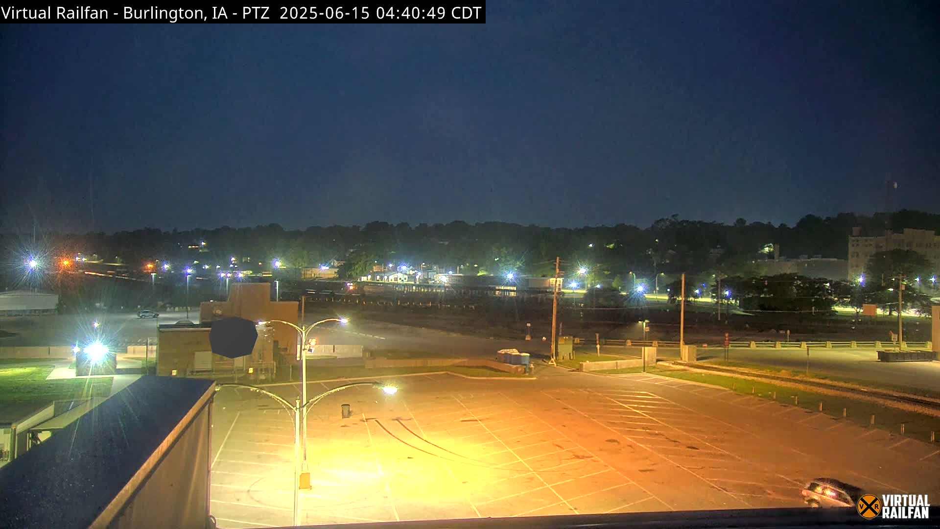 A mostly empty, brightly lit parking lot at night overlooks a tree-lined town under a dark blue sky.