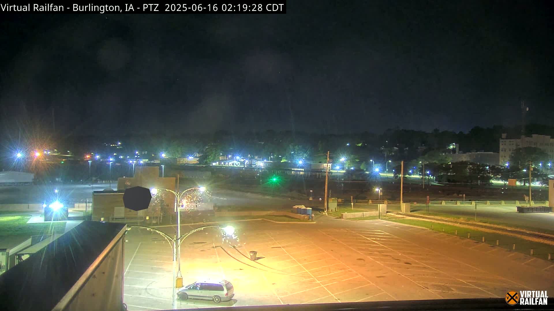 A mostly empty parking lot at night is illuminated by streetlights, with a vehicle parked nearby and a view of a distant town under a clear, dark sky.