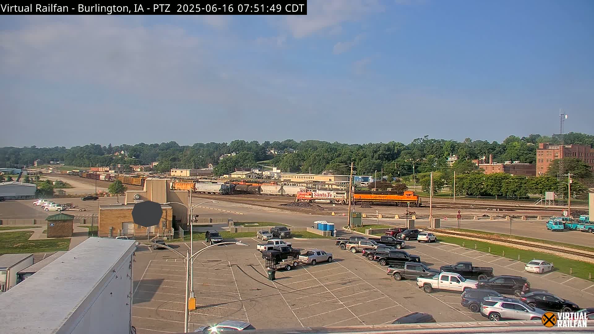 A mostly sunny day reveals a train yard with several locomotives and freight cars, along with a parking lot containing numerous pickup trucks and SUVs.