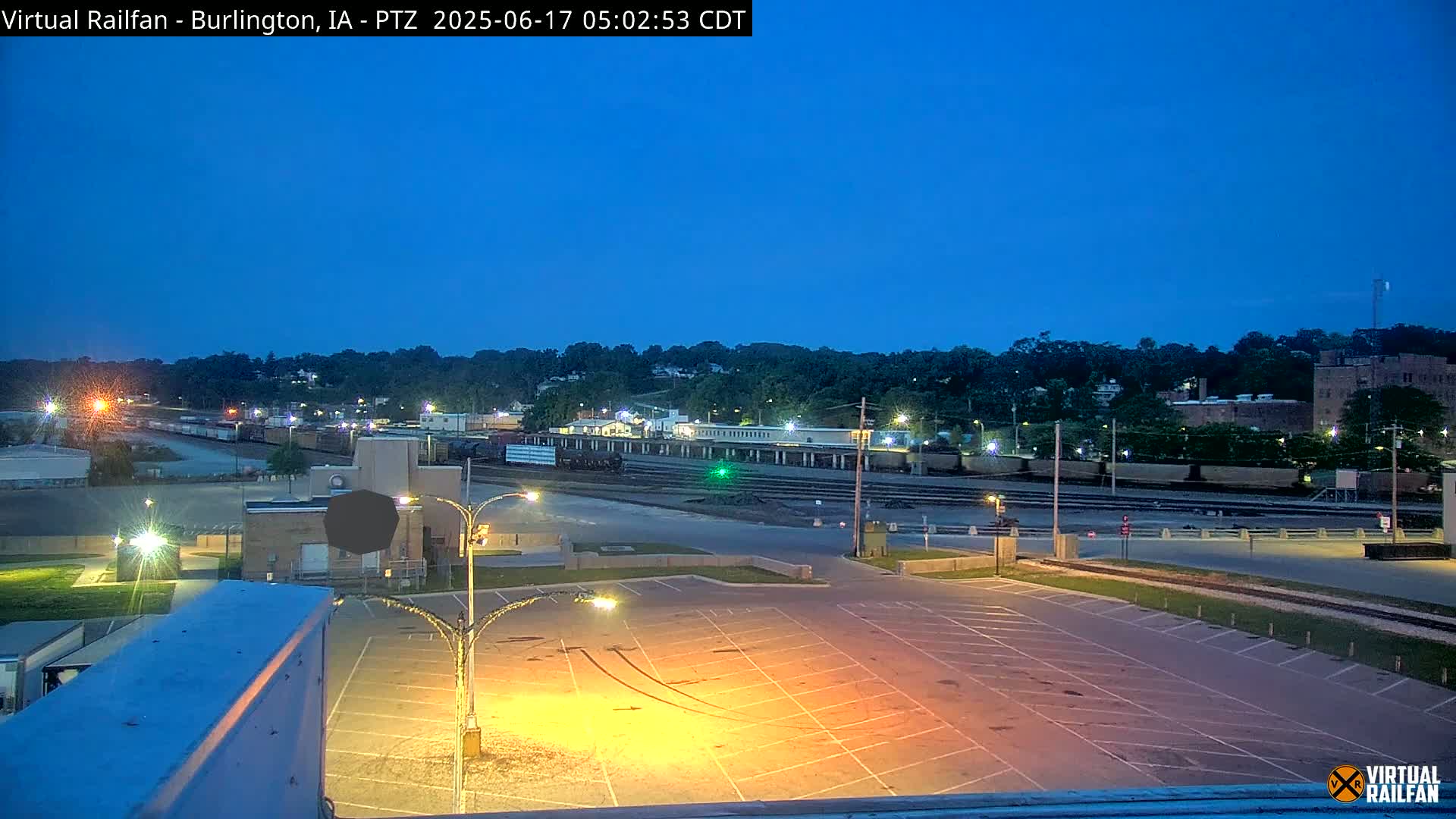 A mostly empty parking lot is illuminated at twilight, overlooking a railroad yard with several train cars and a line of buildings in the background under a clear, dark blue sky.