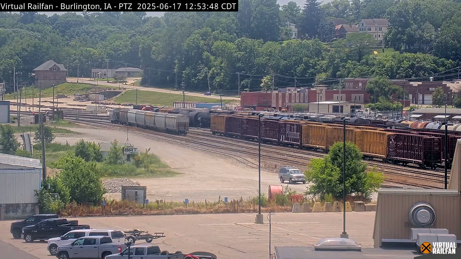 A long freight train sits on railroad tracks in a sunny outdoor setting, with several vehicles parked nearby and a residential area visible in the background.