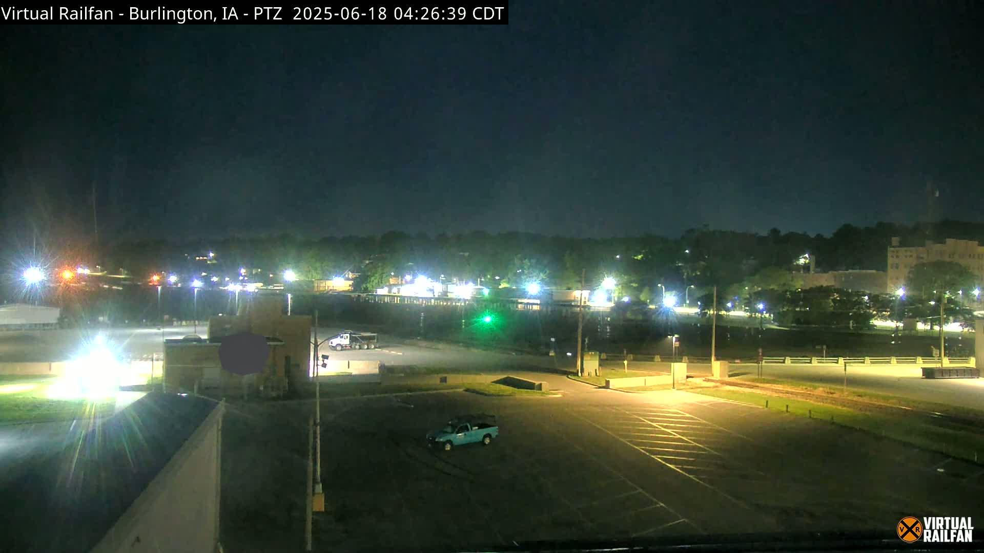 A nighttime view of a mostly empty parking lot with a few vehicles, light posts illuminating the surrounding area, and a treeline in the background under a clear, dark sky.