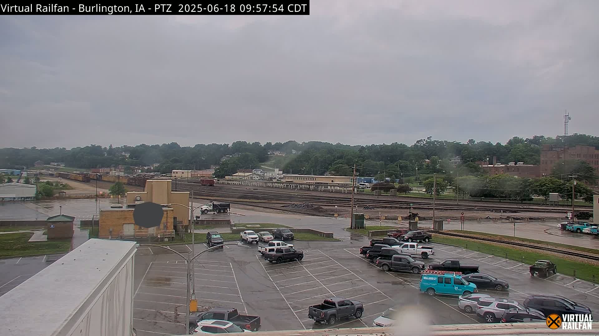 A wet parking lot with several vehicles is shown, overlooking a railroad yard with train cars and a line of buildings under an overcast sky.