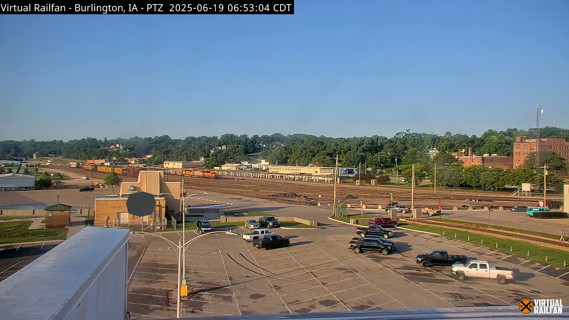 A mostly empty parking lot sits in front of a rail yard with several freight cars, under a clear blue sky.