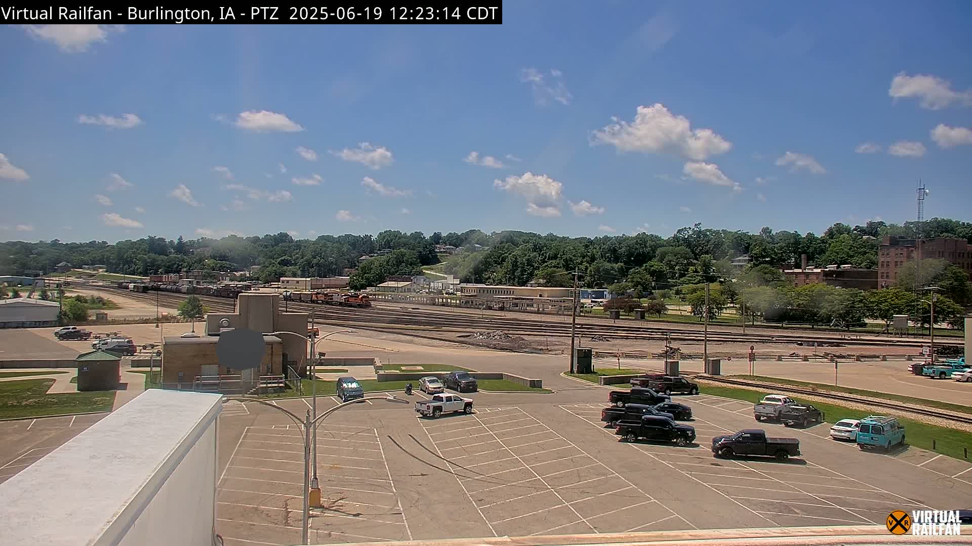 A mostly sunny day reveals a train yard with several parked cars in a lot, and a line of trees and buildings in the background.
