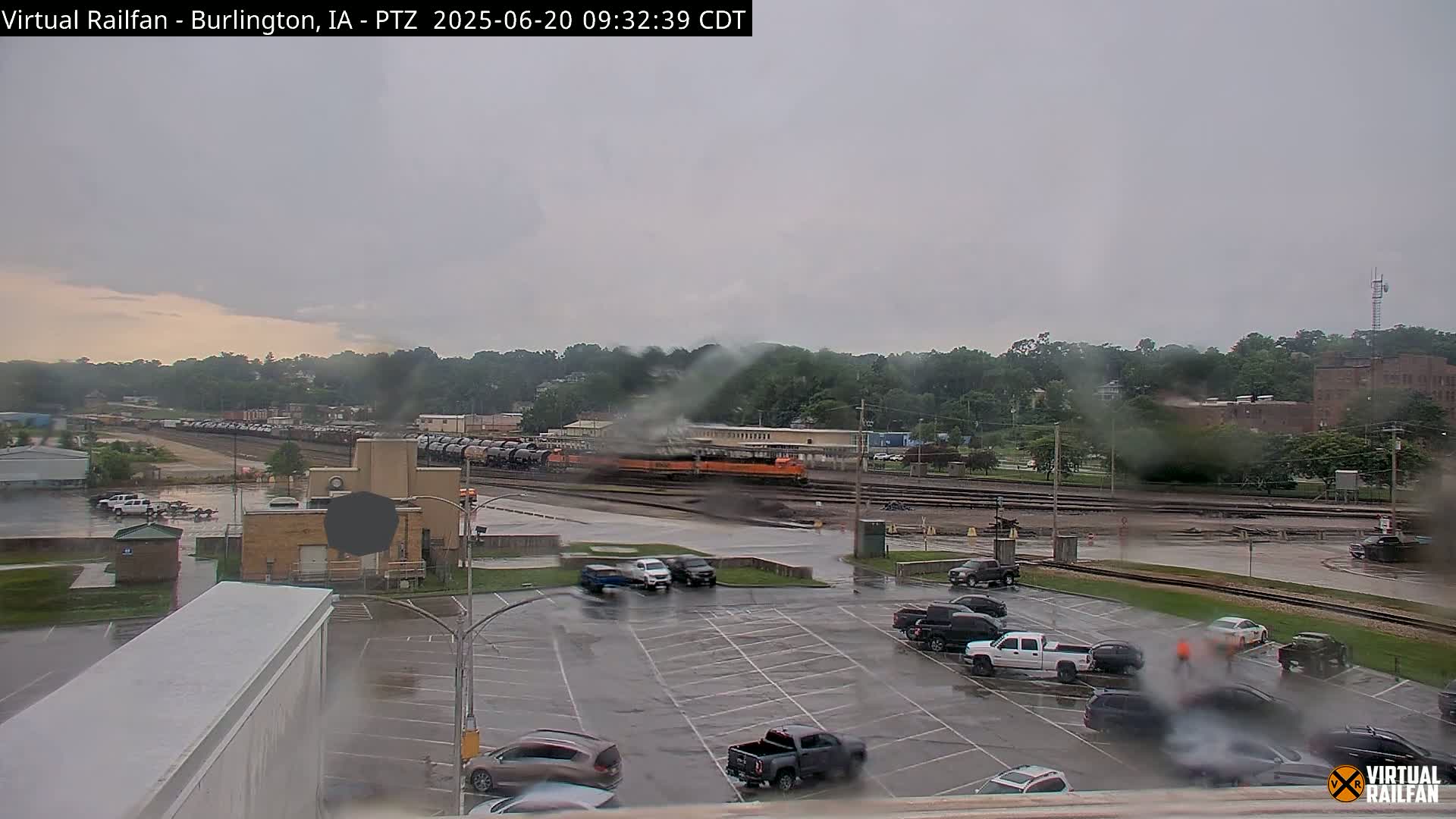 A long freight train passes through a railyard on a rainy day, with cars parked in a lot in the foreground.