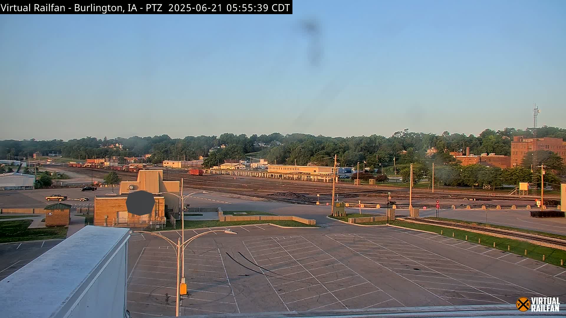 A large empty parking lot overlooks a rail yard with several train cars and industrial buildings under a clear, bright sky.