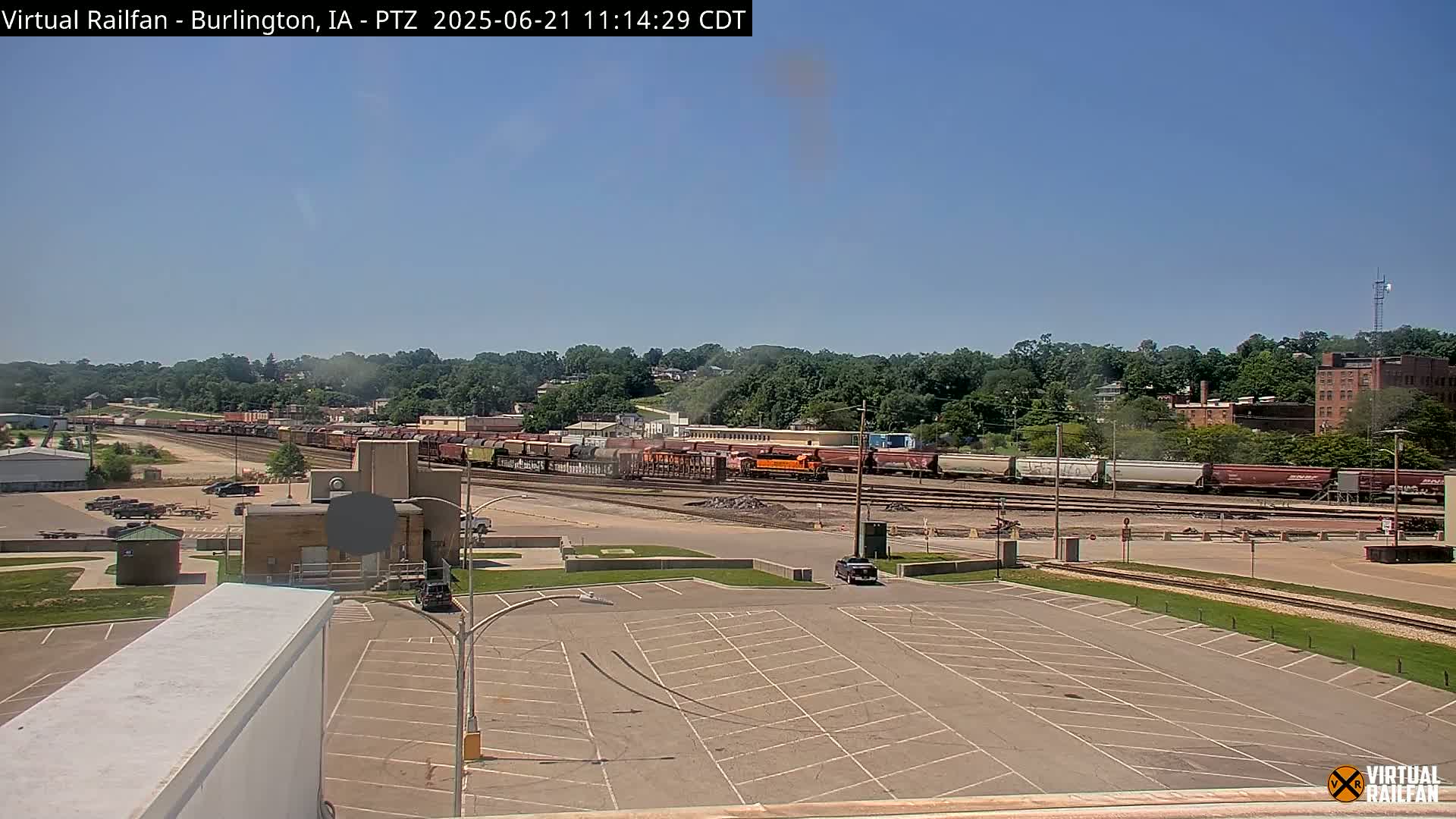 A long freight train is parked in a rail yard on a sunny day, with a large empty parking lot in the foreground.