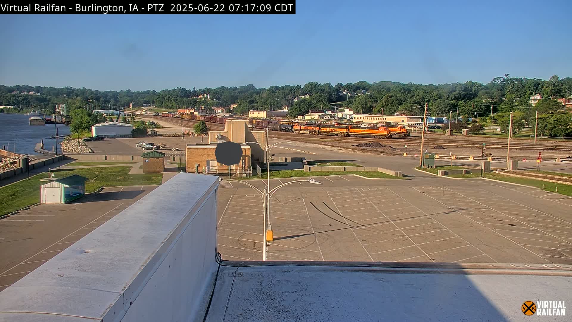 A large, mostly empty parking lot overlooks a train yard with several locomotives and rail cars under a clear, sunny sky near a river.