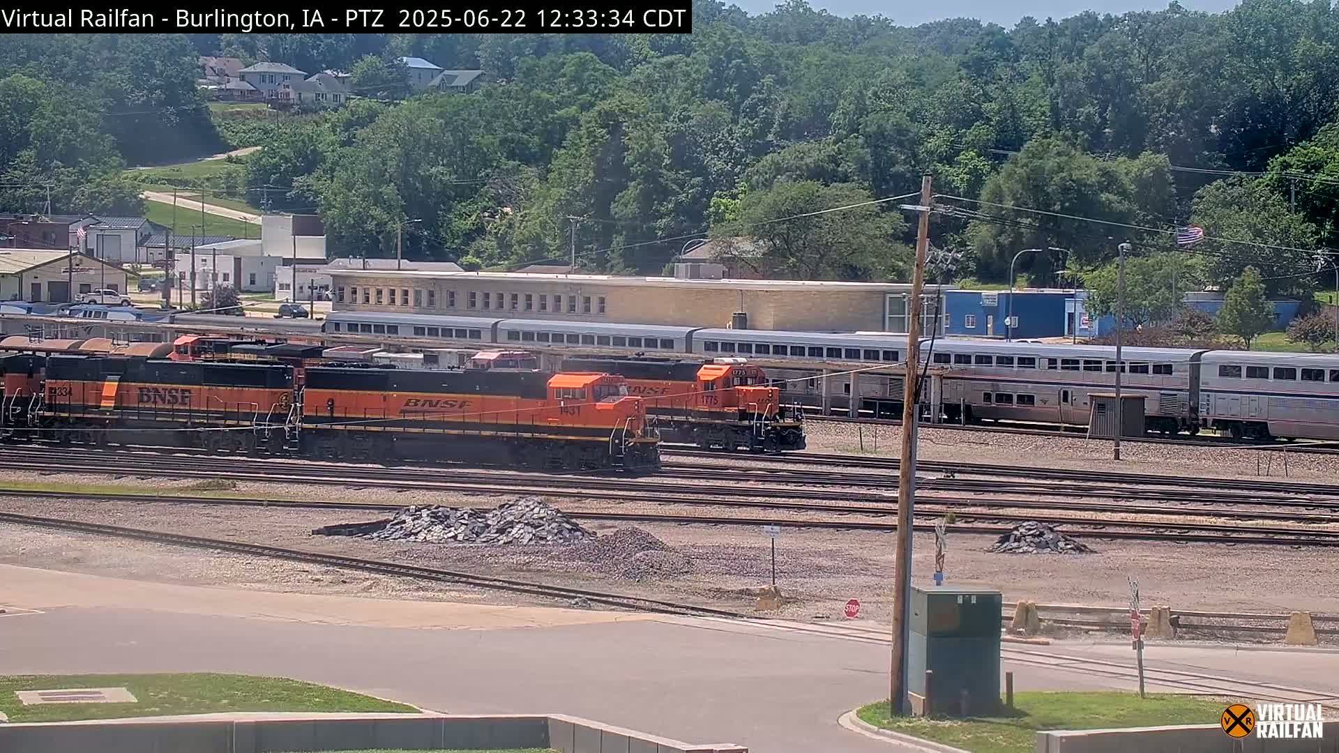 Several orange locomotives and passenger cars sit on a rail yard on a sunny day, with trees and buildings visible in the background.