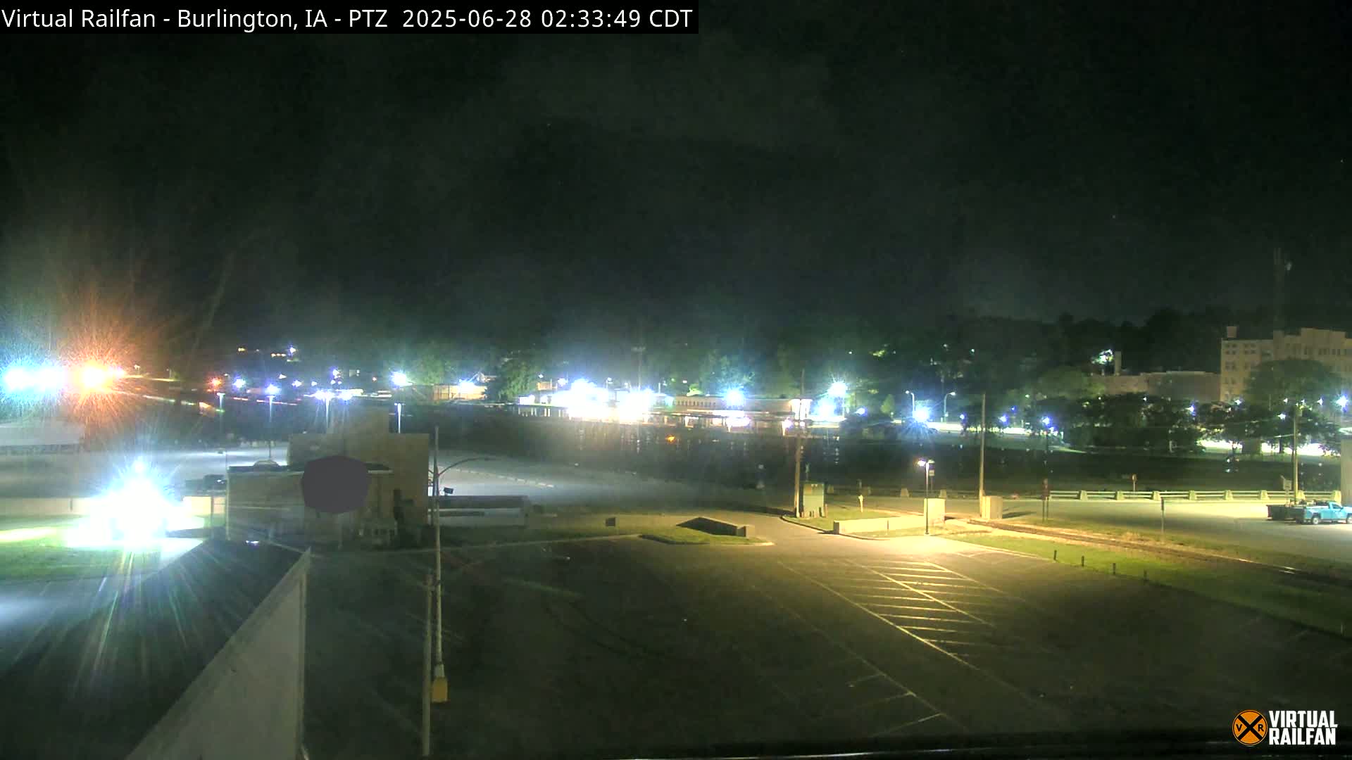 A nighttime view of a mostly empty parking lot, illuminated by streetlights, with buildings and trees visible in the distance under a partly cloudy sky.