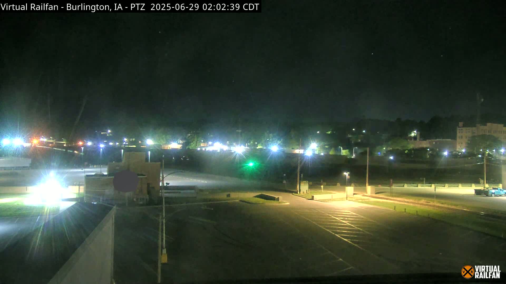 A nighttime view of a mostly empty parking lot and roadway, with lights illuminating buildings and trees in the distance under a clear, dark sky.