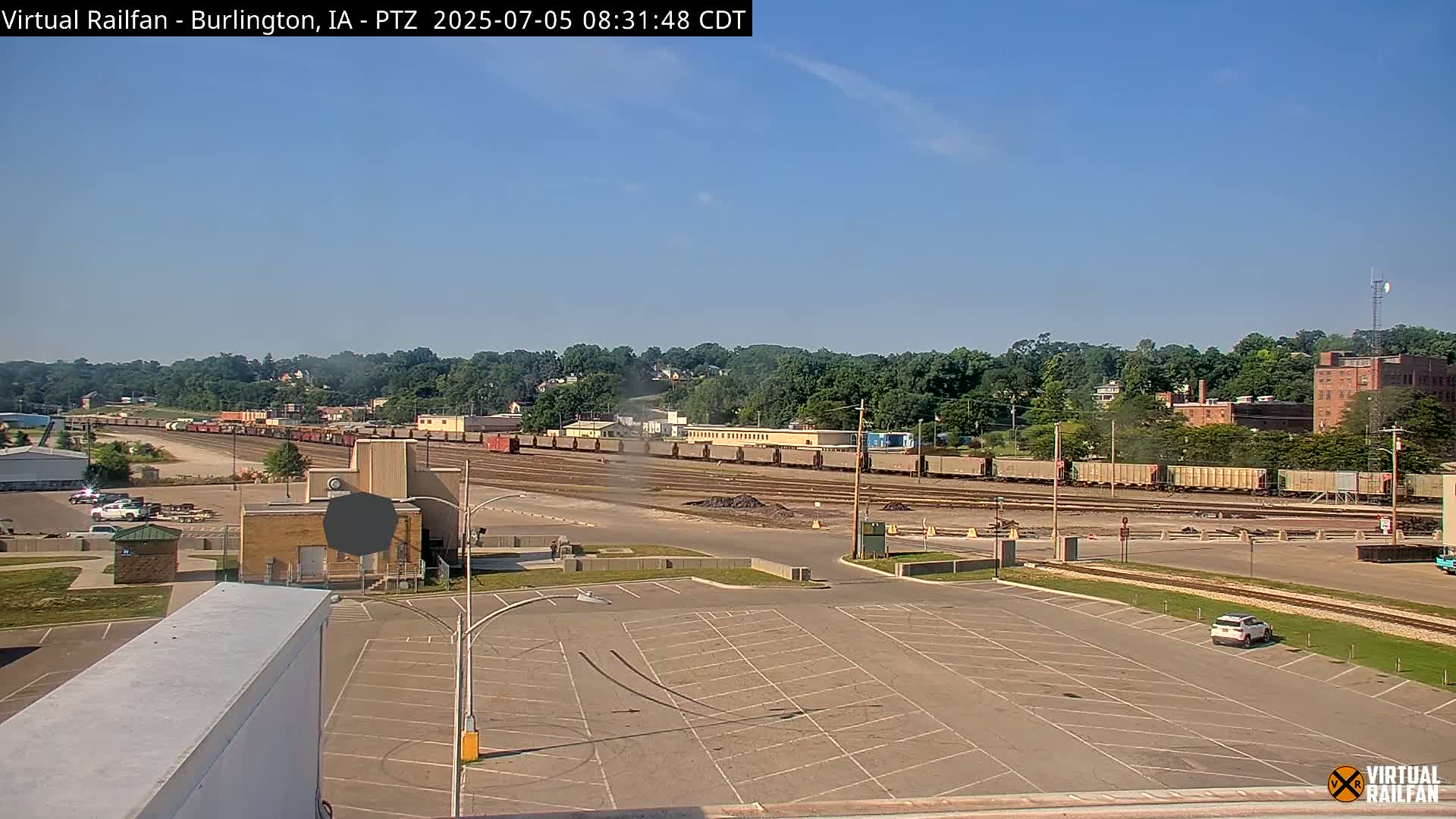 A long freight train sits in a rail yard on a sunny day, adjacent to a large empty parking lot.