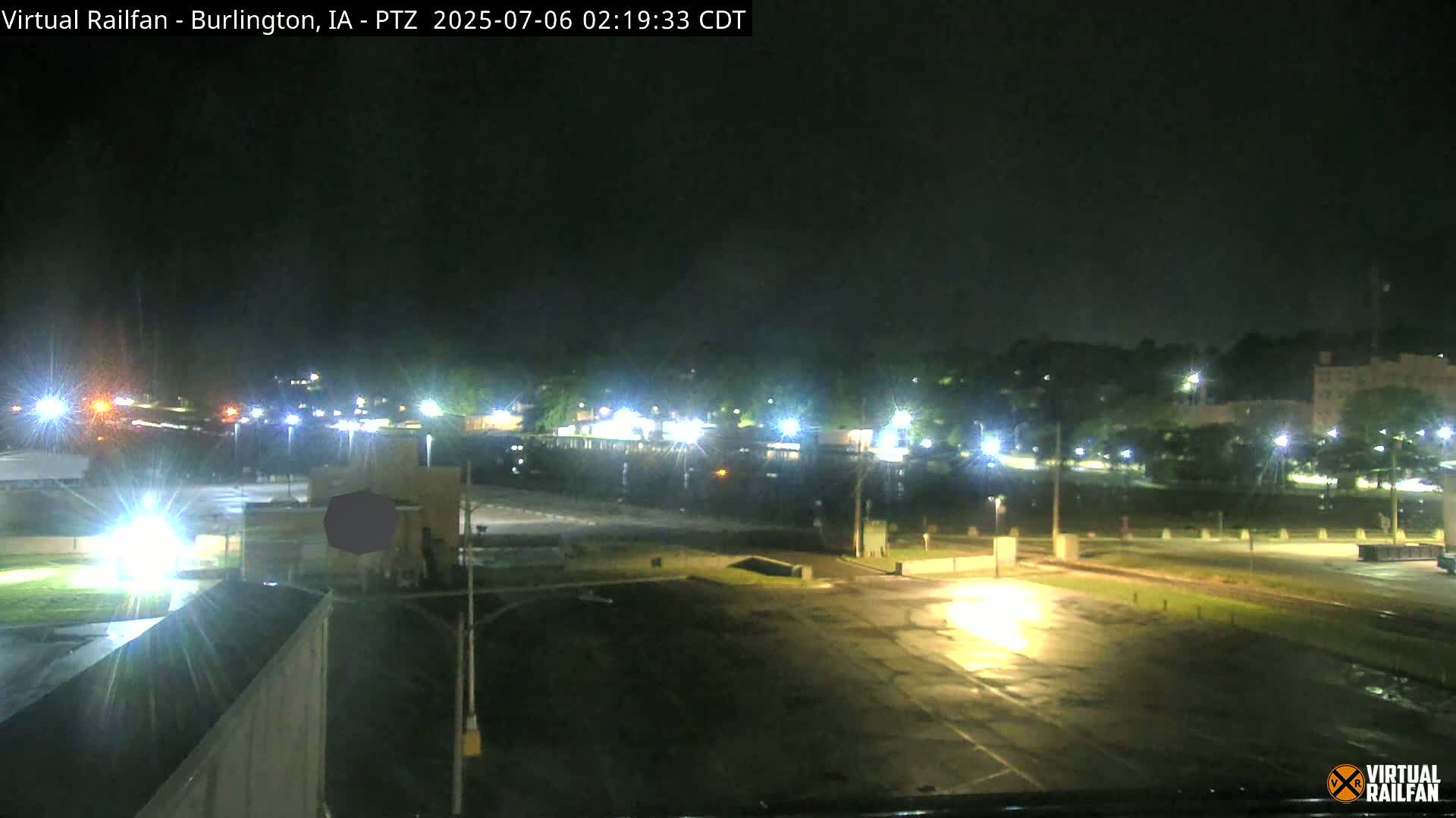 A nighttime view of a mostly empty parking lot and surrounding area, illuminated by streetlights, with a wet surface indicating recent rain.