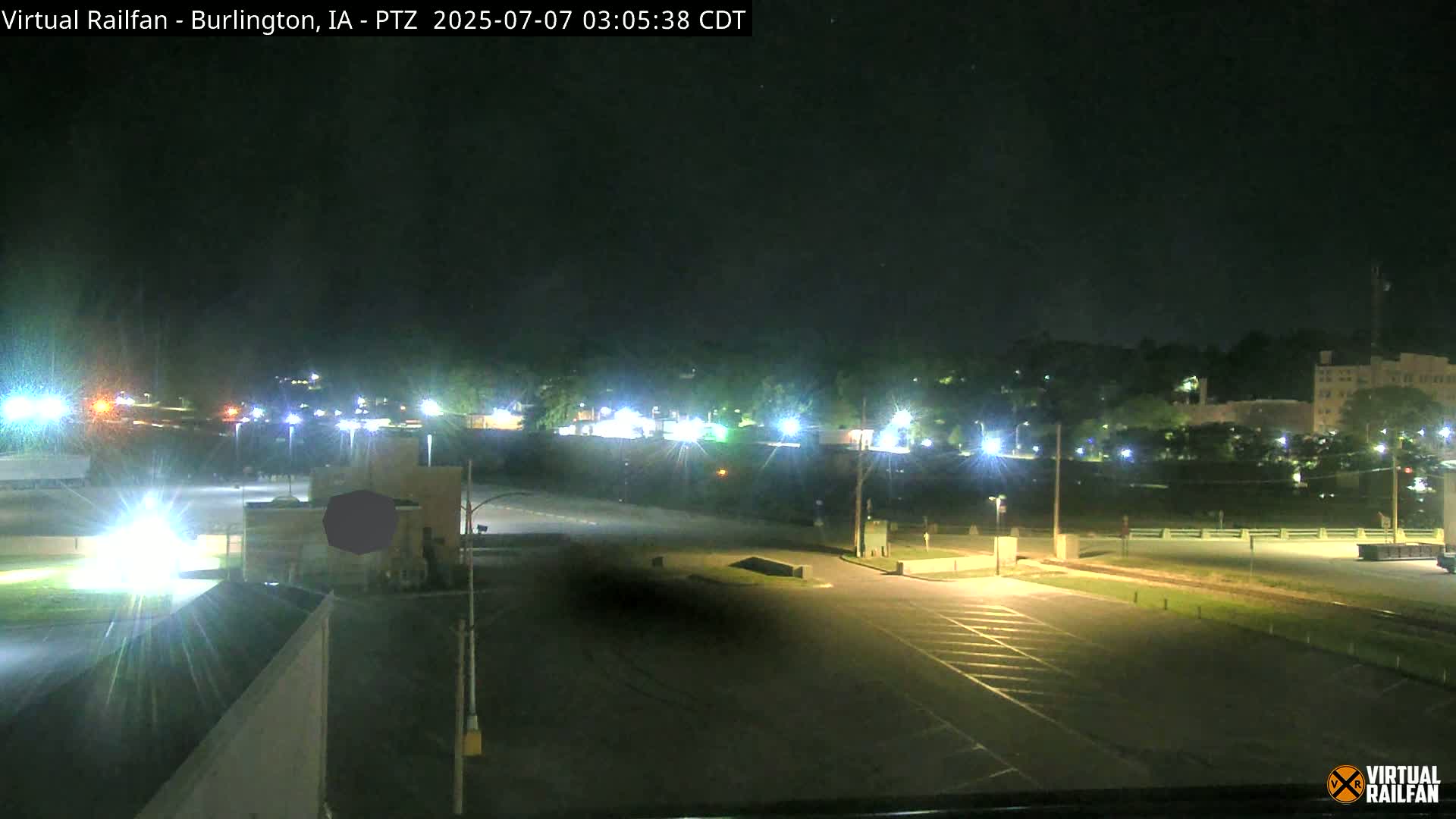 A nighttime, brightly lit, empty parking lot and roadway near a building complex under a clear sky.