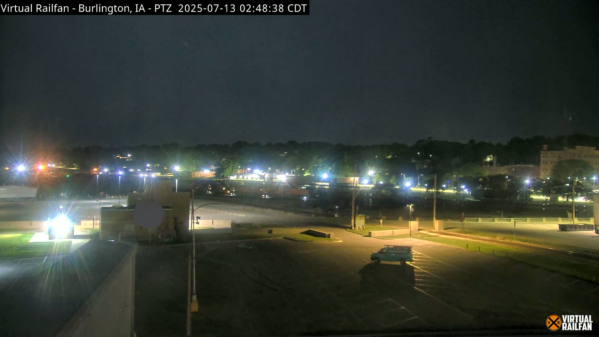 A nighttime view of a mostly empty parking lot, with a few cars visible, and a line of buildings and trees in the background under a dark sky.