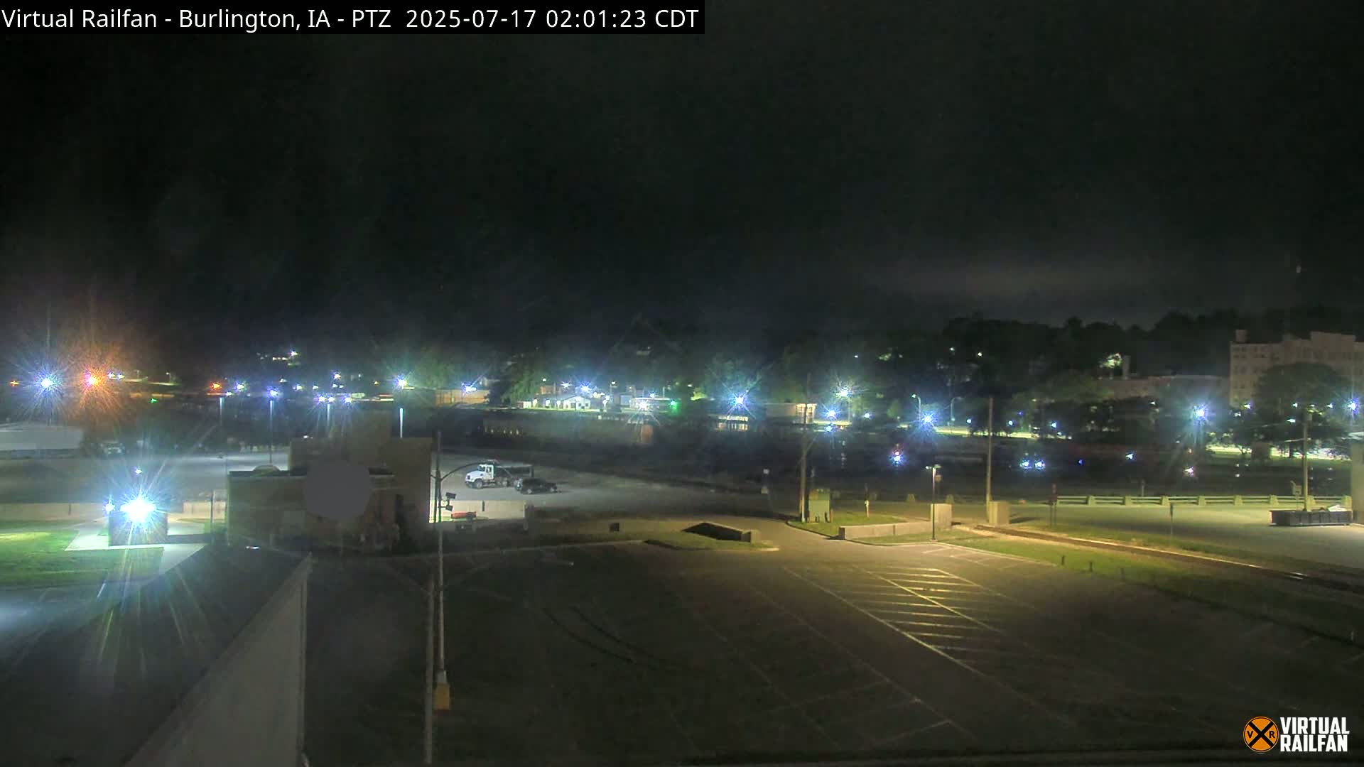 A nighttime view of a mostly empty parking lot next to a road with streetlights illuminating buildings and trees in the distance under a dark sky.