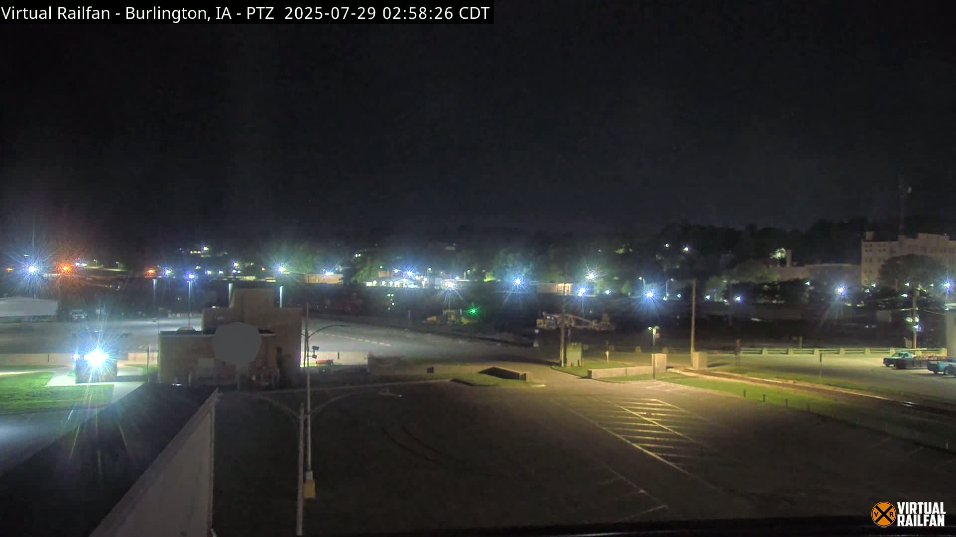 A nighttime view of a mostly empty parking lot and surrounding area, illuminated by streetlights, with a line of trees and buildings in the distance under a clear, dark sky.