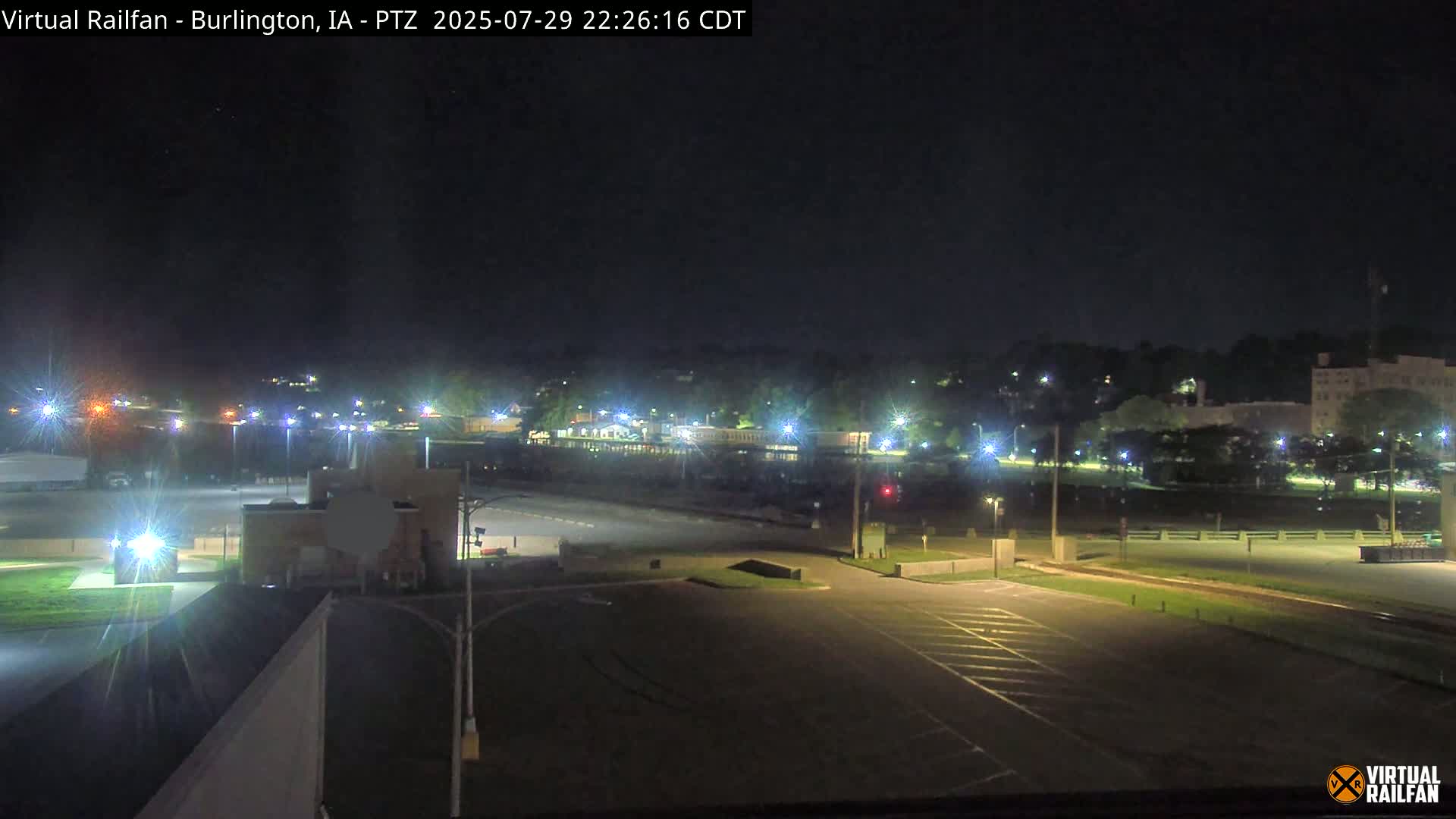 A nighttime view of a mostly empty parking lot and surrounding area, illuminated by streetlights, with a line of trees and buildings in the distance under a clear, dark sky.
