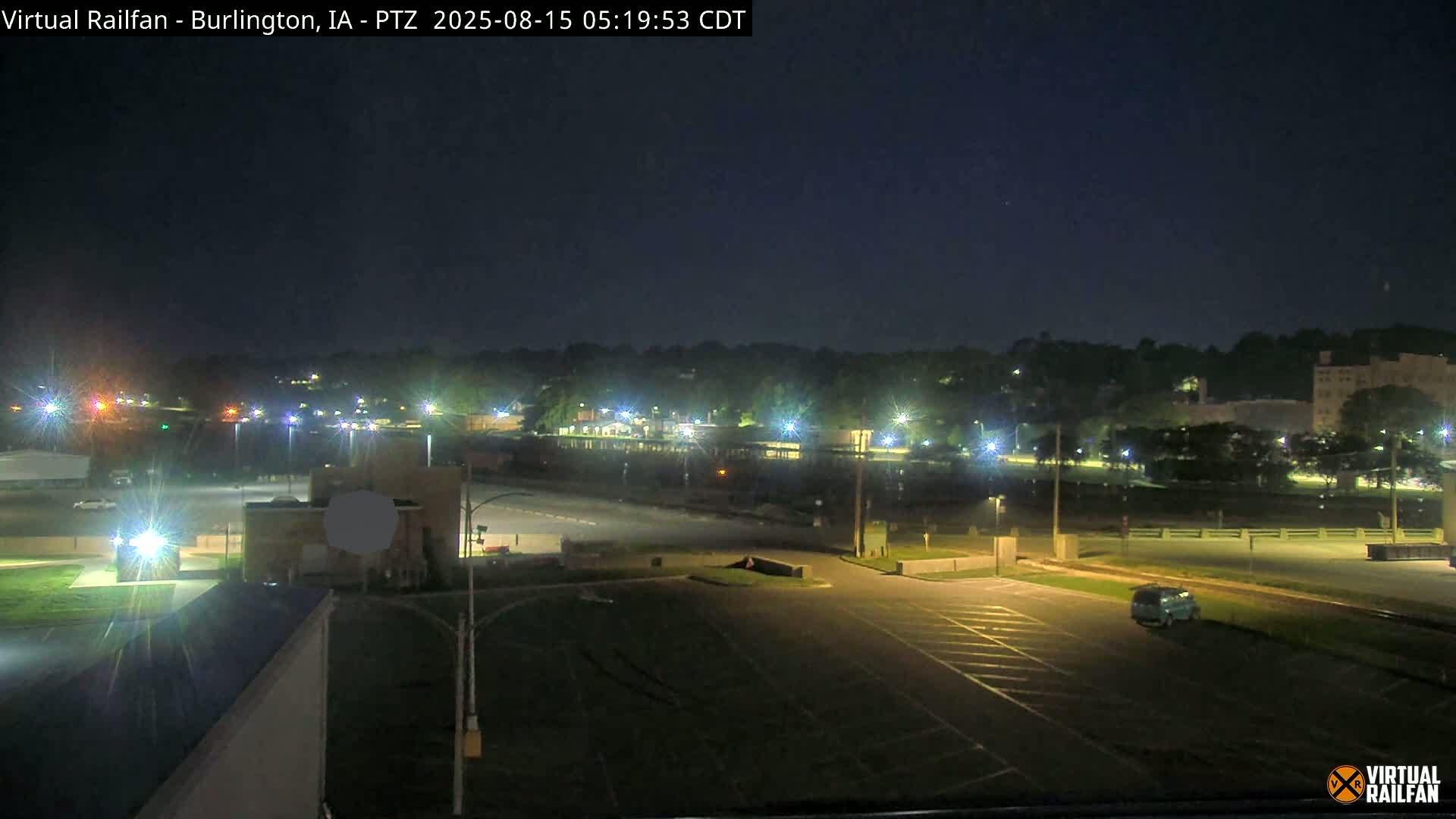 A nighttime view of a mostly empty parking lot and surrounding area, illuminated by streetlights, with a line of trees and buildings in the distance under a clear, dark sky.