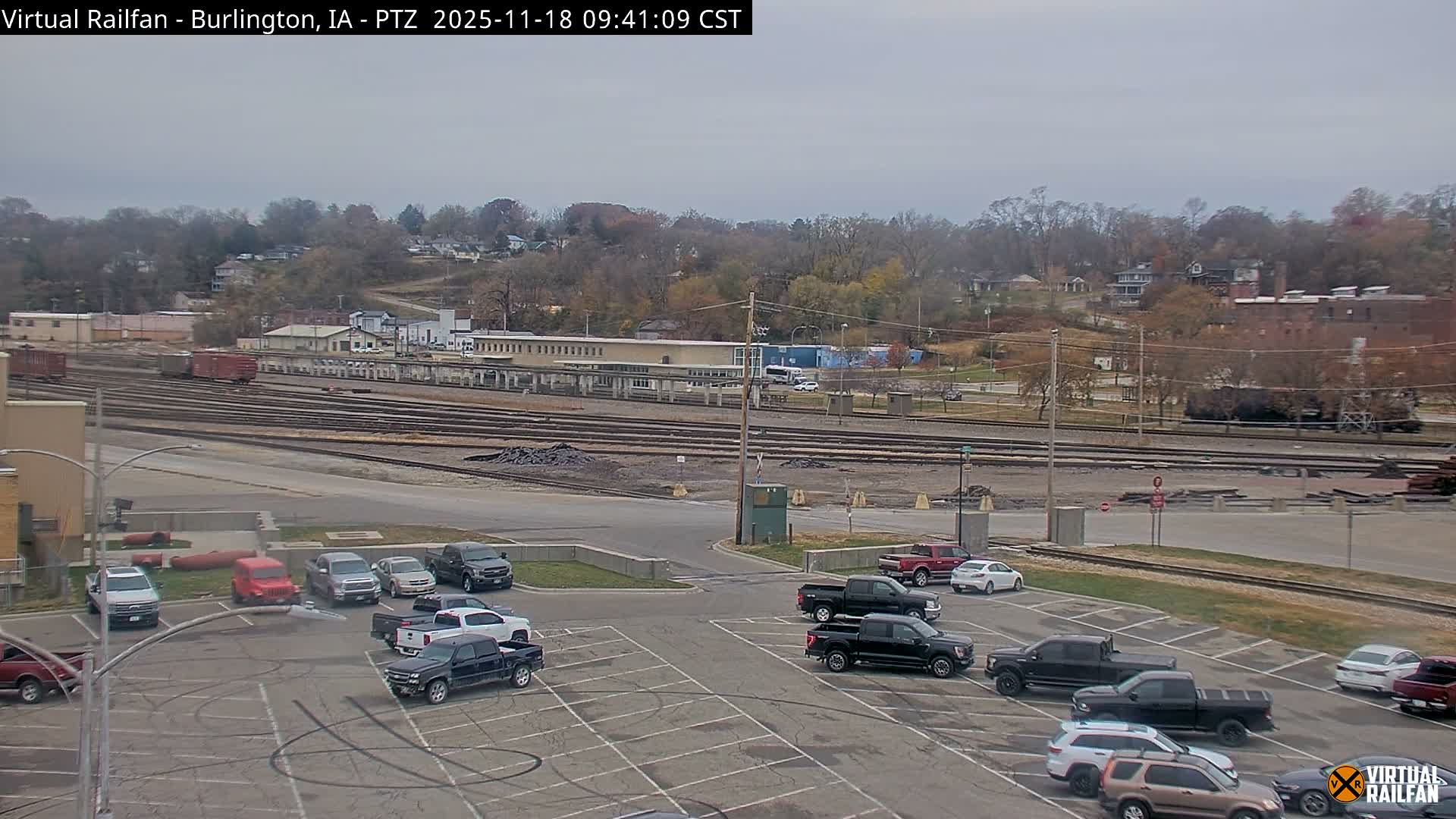 A nighttime view of a mostly empty parking lot and surrounding area, illuminated by streetlights, with a line of trees and buildings in the distance under a clear, dark sky.