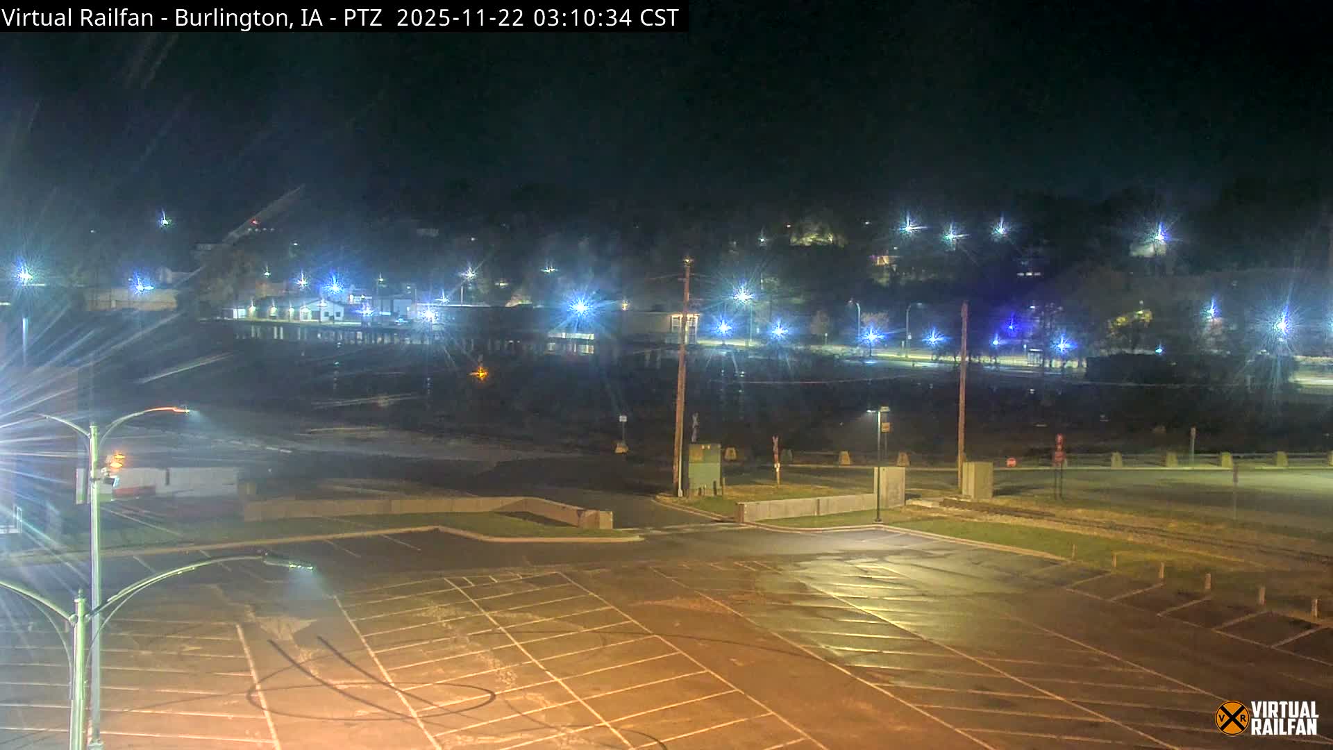 A brightly lit, wet parking lot and roadway are visible at night, with railway tracks in the midground and numerous distant city lights twinkling across a dark river or valley, indicating damp conditions.