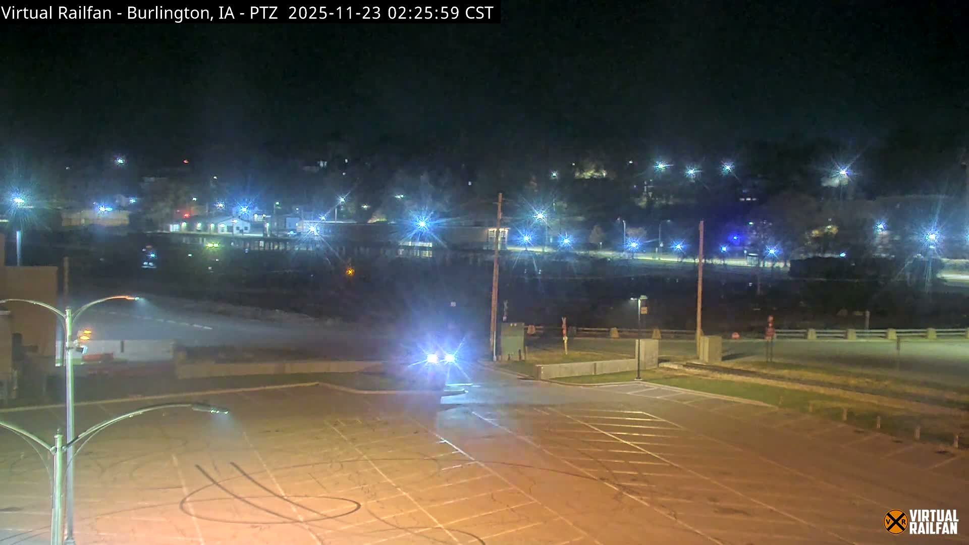 Under a clear night sky, a bright vehicle with glaring headlights occupies the foreground of a paved area, while numerous streetlights and building lights illuminate a distant urban landscape across a lower-lying dark expanse.