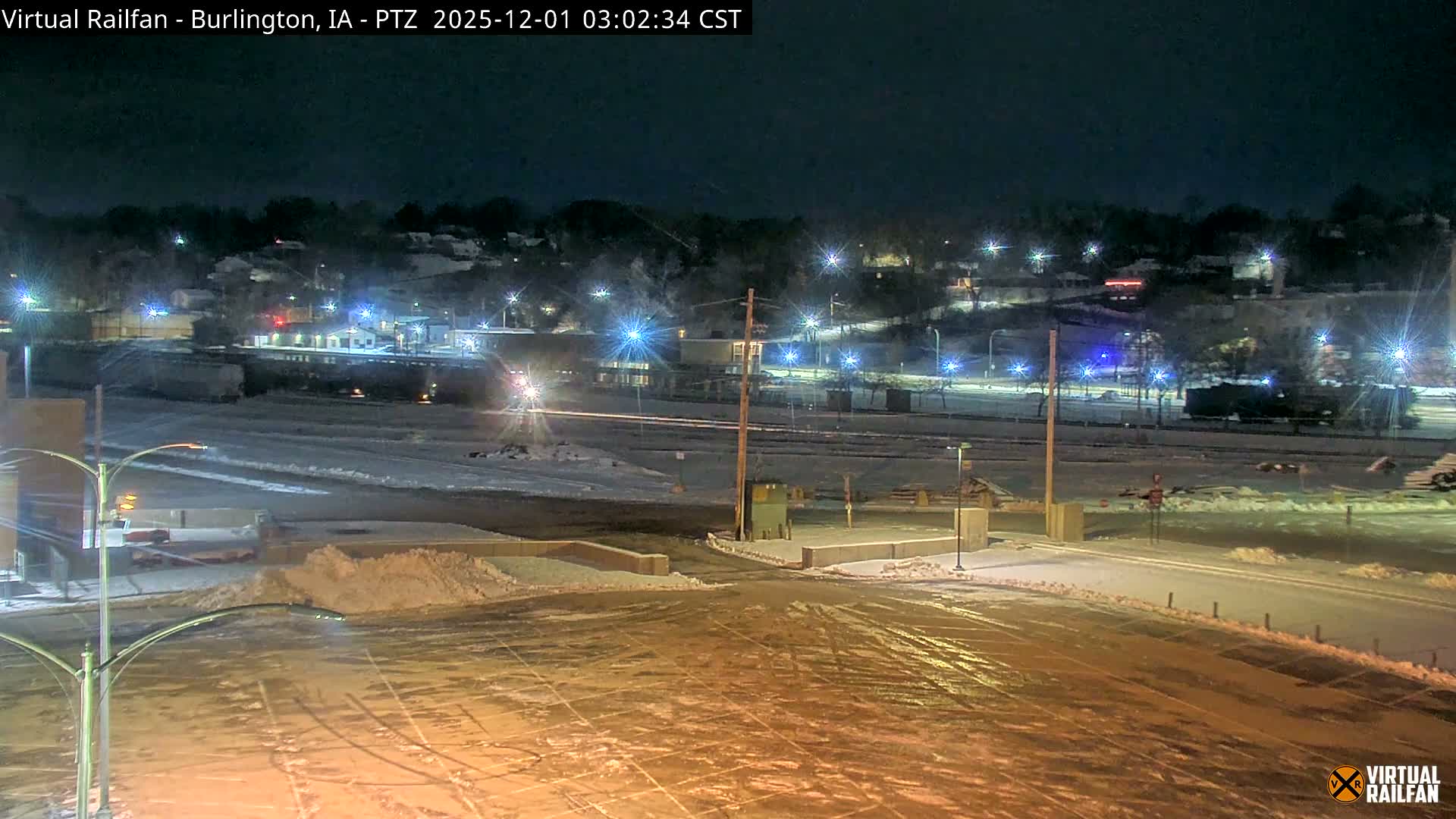 The image displays a snow-covered railroad yard and adjacent roads at night, brightly illuminated by numerous streetlights and building lights under clear, cold conditions.