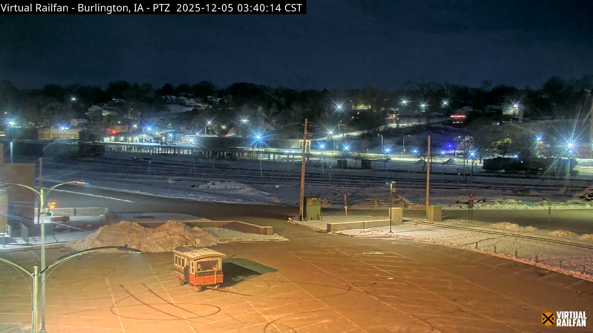 A winter night scene reveals a brightly lit, snow-dusted parking lot with a small, old-fashioned wagon in the foreground, set against numerous illuminated train tracks and distant town lights under a dark sky.