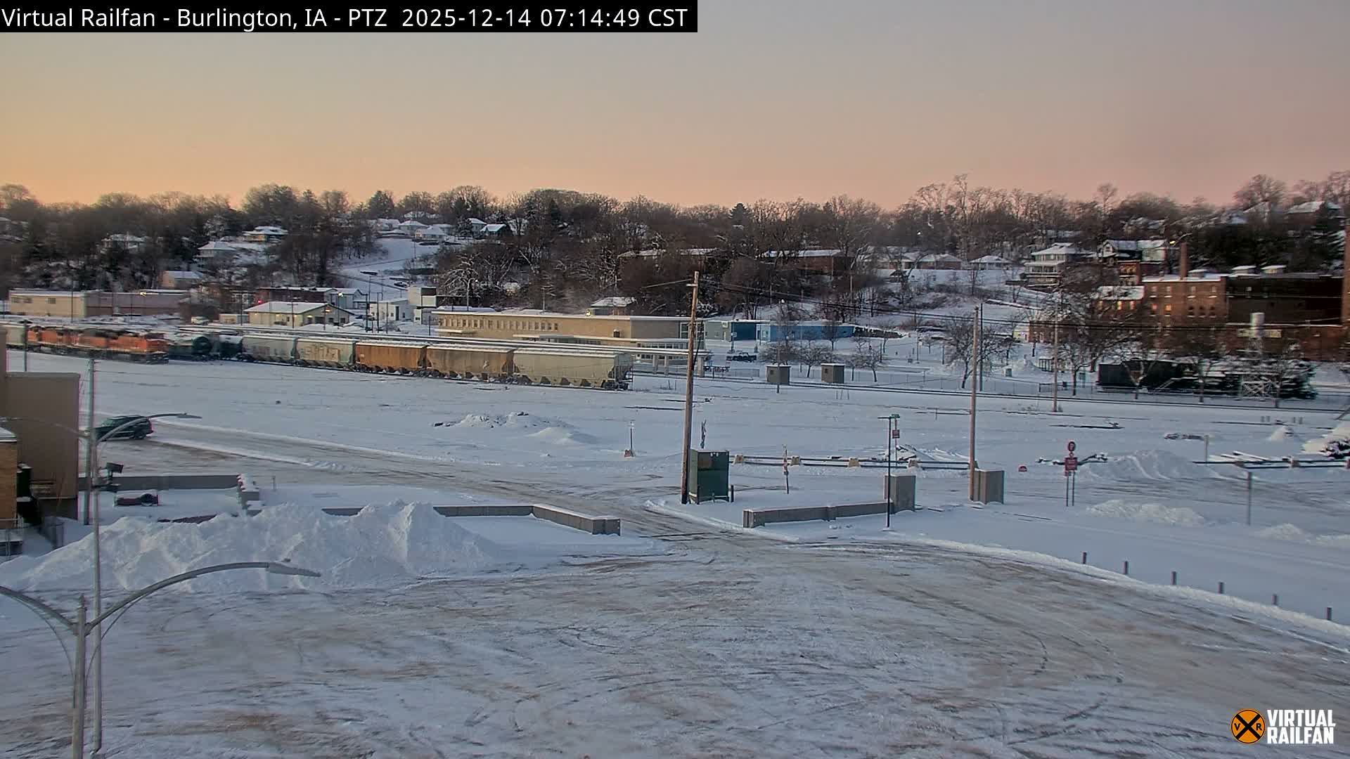 A winter night scene reveals a brightly lit, snow-dusted parking lot with a small, old-fashioned wagon in the foreground, set against numerous illuminated train tracks and distant town lights under a dark sky.