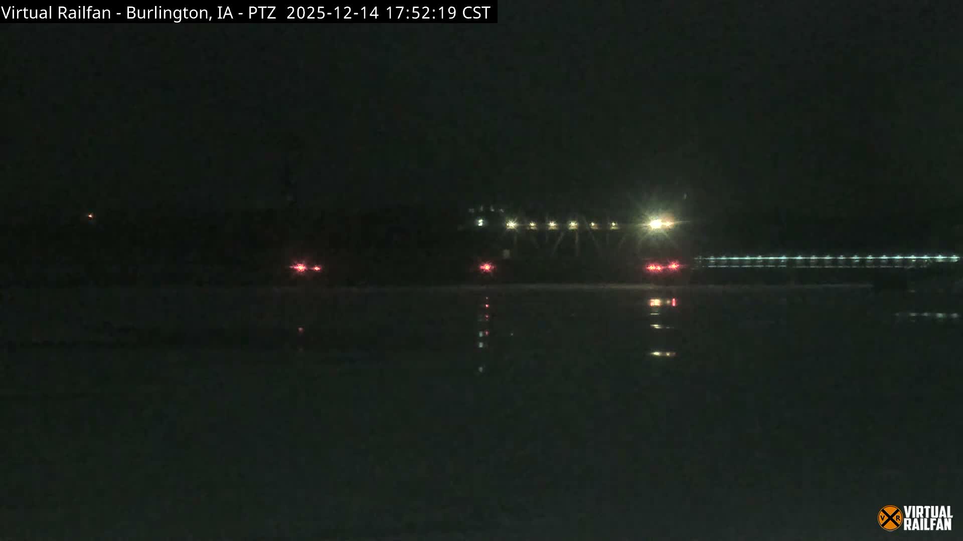 A winter night scene reveals a brightly lit, snow-dusted parking lot with a small, old-fashioned wagon in the foreground, set against numerous illuminated train tracks and distant town lights under a dark sky.