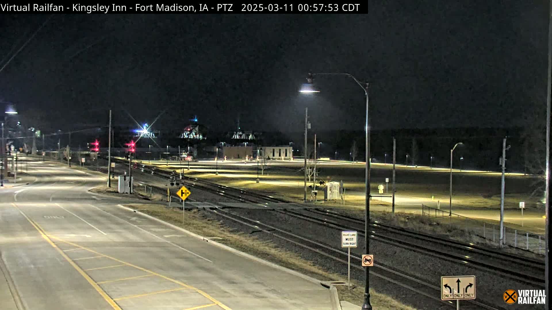 A nighttime view of a road intersection near railroad tracks, with streetlights illuminating a mostly empty parking lot and surrounding area.
