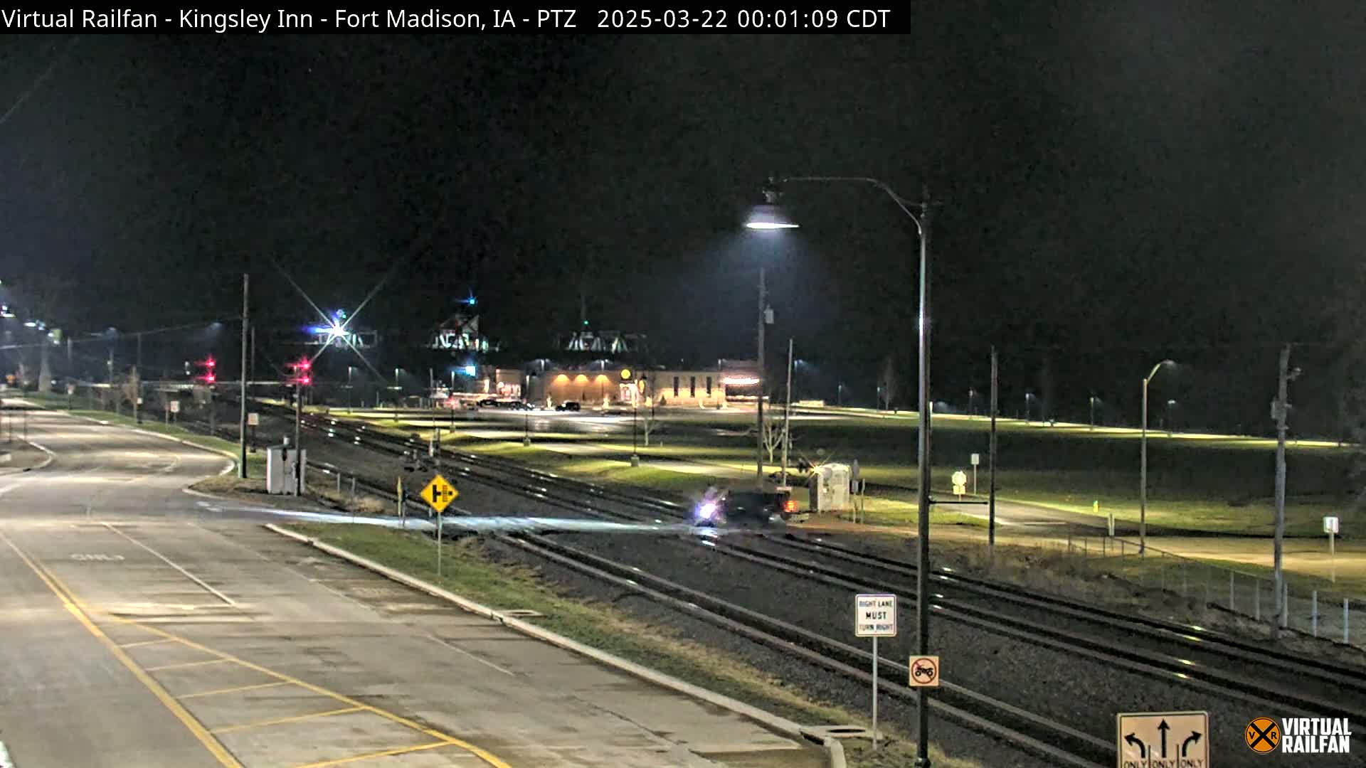 A nighttime, low-angle view of a roadway intersecting railroad tracks, with a vehicle crossing the tracks and a building in the background under clear skies.