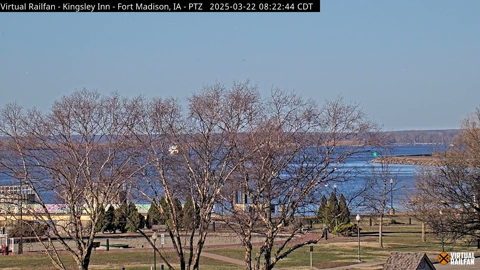 A calm, blue river is seen through bare trees in a park-like setting under a clear, sunny sky.
