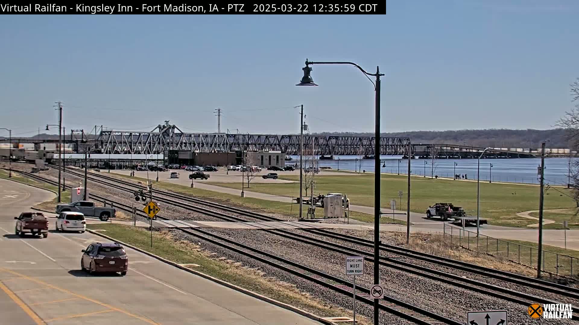 A sunny day shows a view of railroad tracks, roadways, a river with a bridge in the background, and several vehicles.