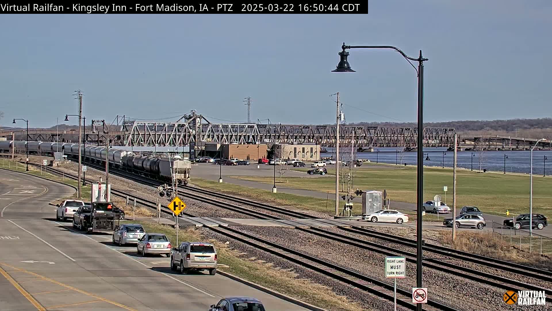 A long freight train passes by a river and bridge on a sunny day, with cars driving on a road next to a railway crossing.