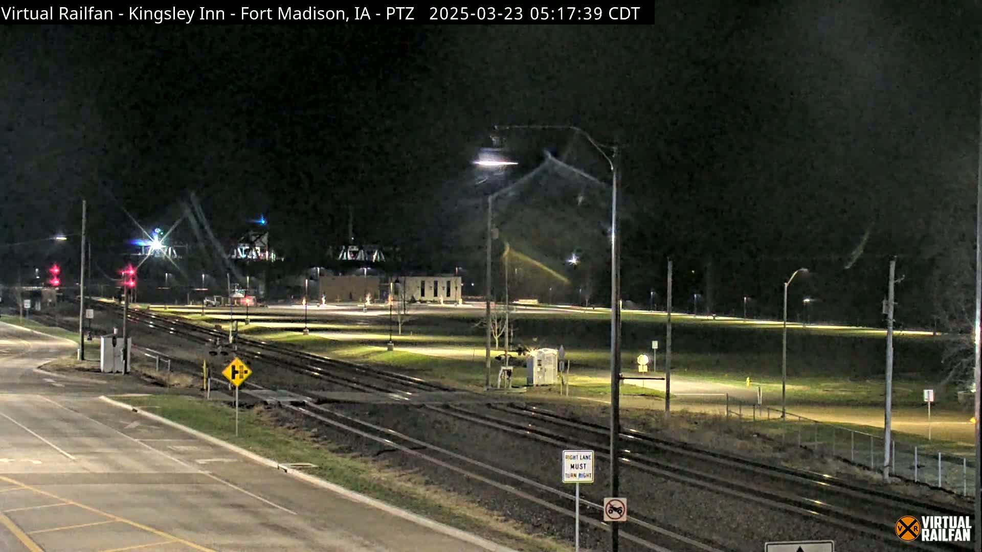 A nighttime view of railroad tracks intersecting with a road, featuring streetlights illuminating a mostly empty parking lot and grassy areas.