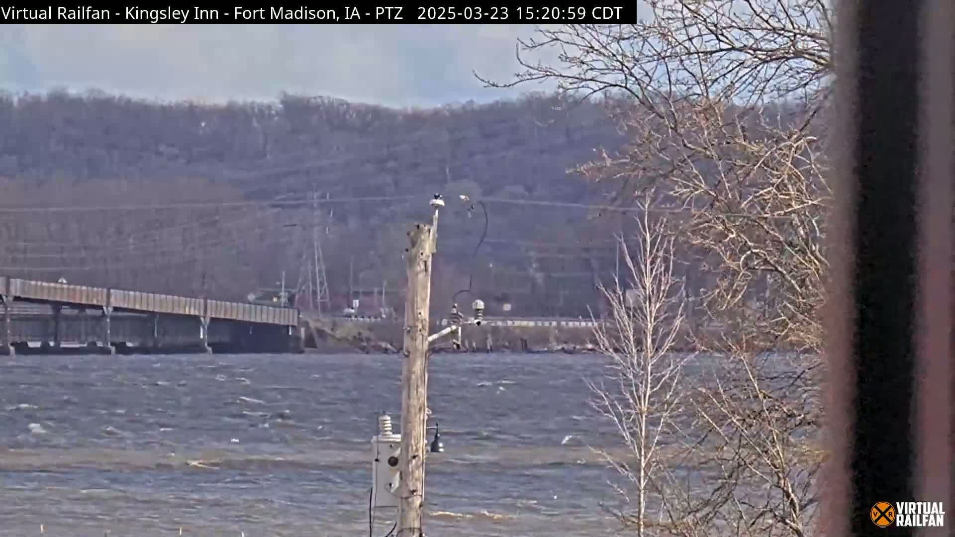 A windy day shows a choppy river with a bridge in the background, a utility pole with equipment in the foreground, and bare trees along the shoreline.