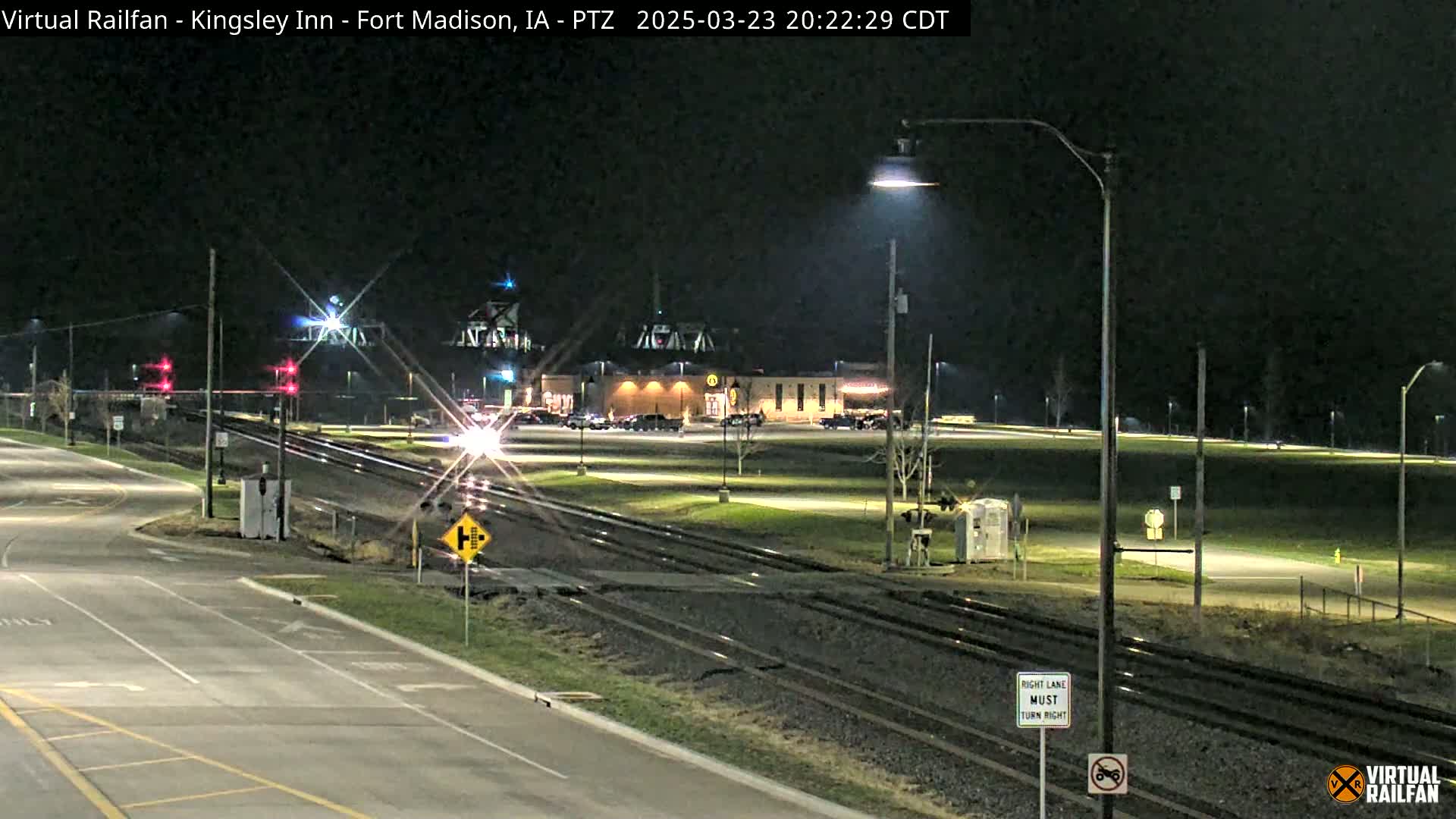 A nighttime view of a road and railroad crossing near a building with cars parked in its lot, under clear skies.