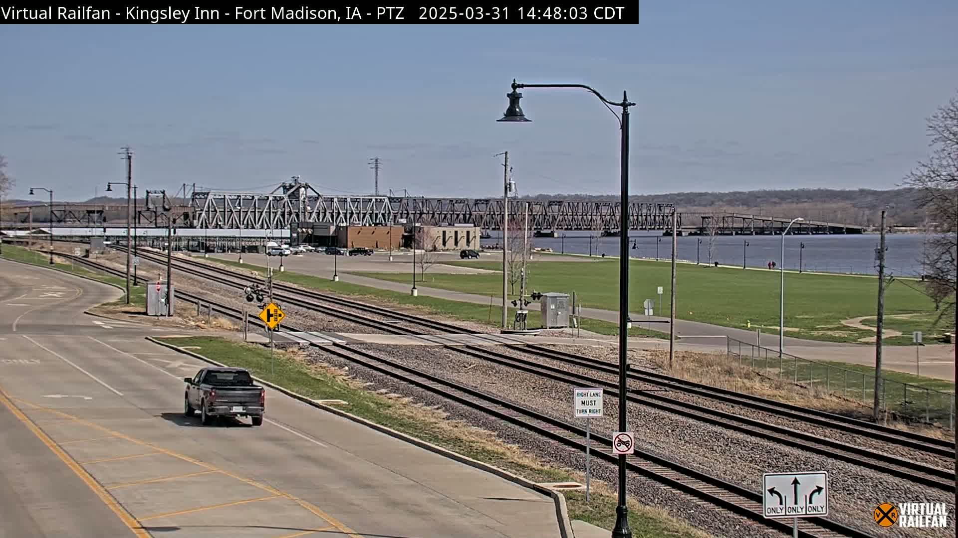 A pickup truck drives on a road next to train tracks, with a metal bridge and river in the background on a sunny day.